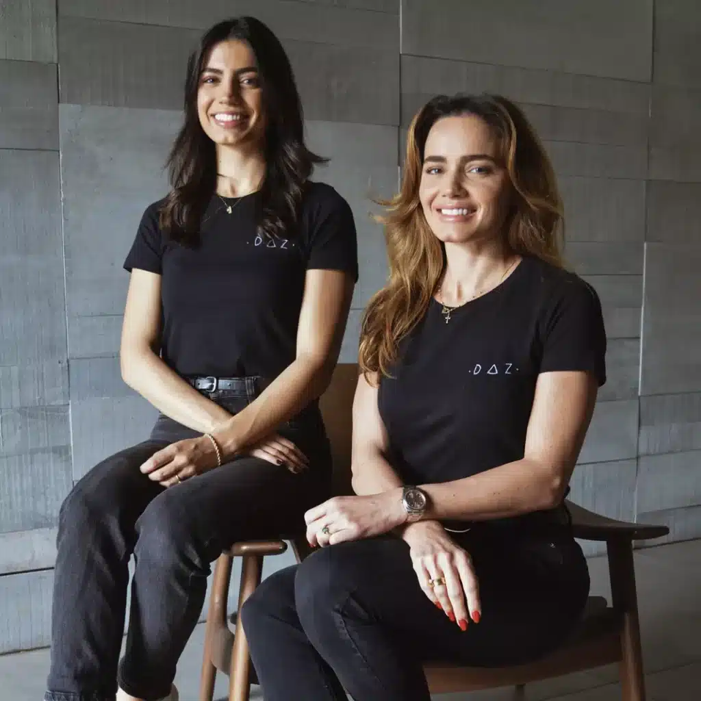 Foto mostra as duas mulheres sentadas, uma ao lado da outra, vestindo roupas escuras